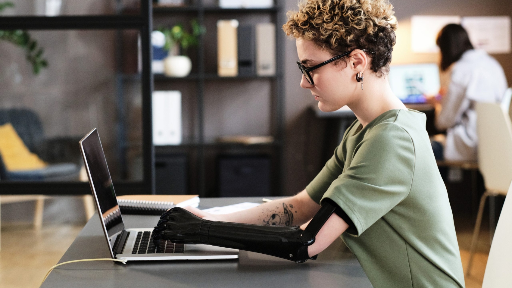 Young woman with prosthetic arm typing on laptop while sitting at office desk
